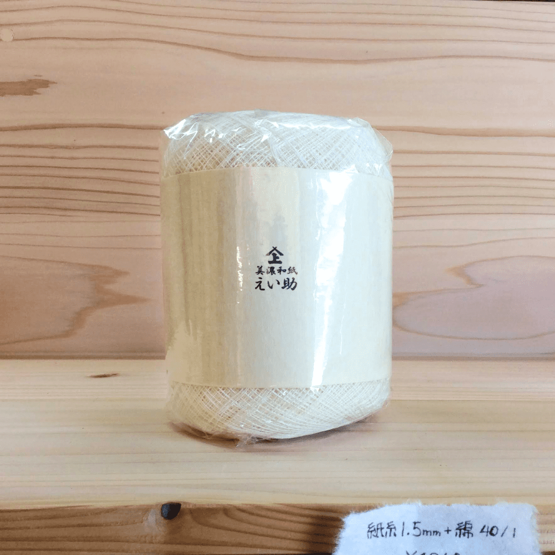 ナチュラルな木材の背景に、紙糸が玉の状態で置かれています。商品真ん中には店名がはいった商品帯が横にまかれています。原料内訳は美濃和紙、綿です。商品名は紙糸1.5mm+綿40/1です。下記は関連するキーワードになります。紙糸 和紙糸 和紙素材 美濃和紙 オリジナル ニット 編み物 小物づくり アクセサリー作り クラフト ハンドメイド 刺繍 アート 作品 素材 自然素材 近くで和紙が売ってるところ 松久永助紙店 オンラインショップ