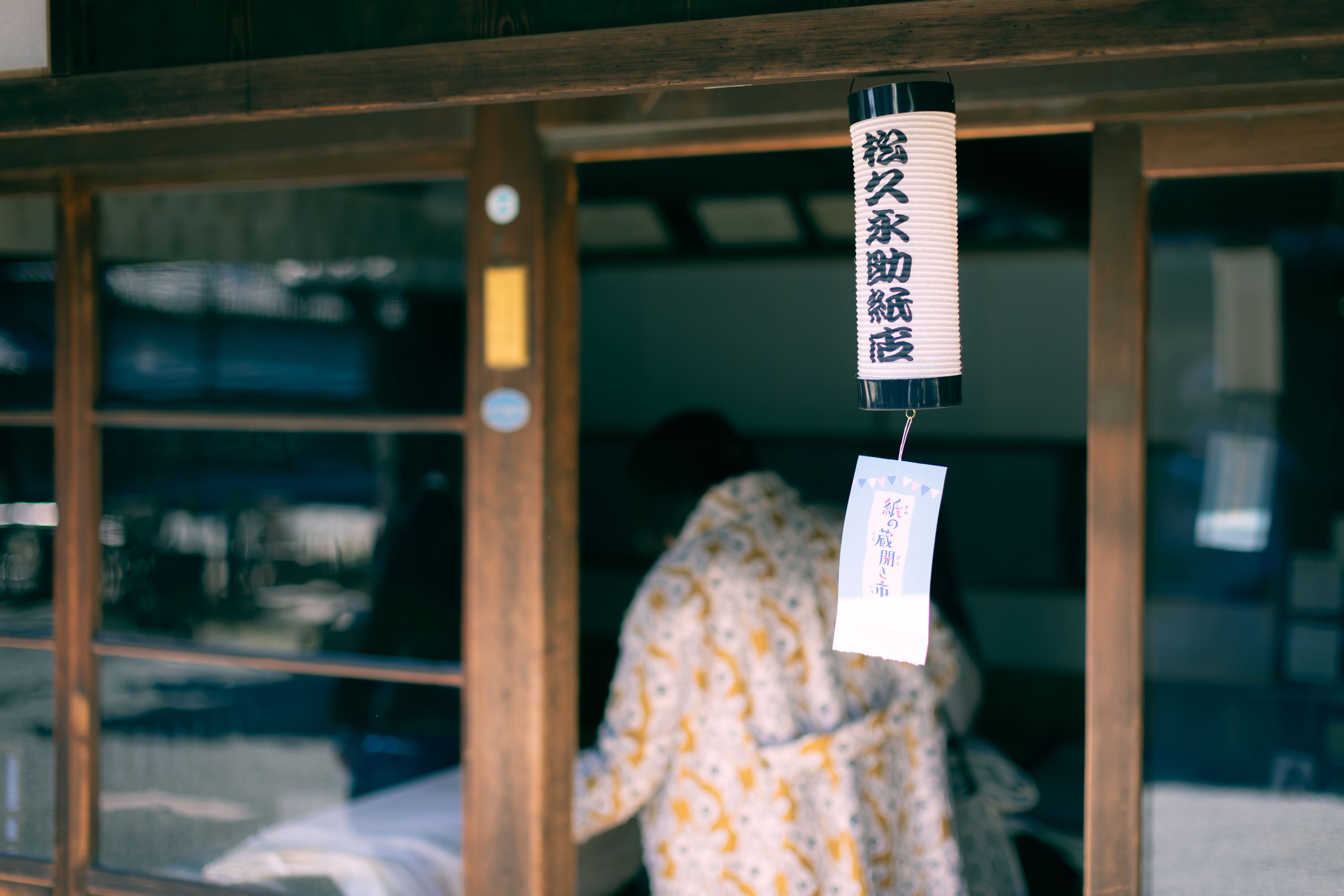 松久永助紙店の現店舗向かいにある、帳場に入っていく女性の後ろ姿がうつっています。軒先には店名が入った小さな提灯、その下に「紙の蔵開き市」と記載された紙が吊り下げられています。この紙の蔵開き市は2023年2月に開催されました。下記は関連するキーワードになります。うだつの上がる町並み 老舗 和紙 美濃和紙 和紙問屋 蔵開き 蔵出し 紙の蔵開き市 ヴィンテージ品 デッドストック 貴重 レア 在庫ある限り 美濃 岐阜 近くで和紙が売ってるところ 松久永助紙店