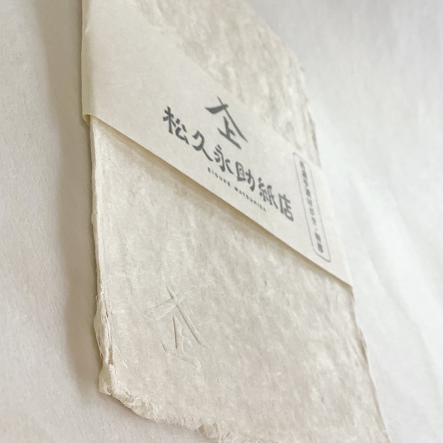 白い背景、「特厚美濃和紙手漉はがき」の表面を、厚みが分かるよう、やや傾けて撮影した写真です。商品真ん中には、店名、商品名を記載した商品帯が横方向にまかれています。左下にはエンボス加工されたロゴマークが見えます。松久永助紙店オンラインショップでは他にも次のような和紙商品を取り扱っています。和紙(楮紙/こうぞ紙・マニラ麻・落水紙・本美濃紙/那須楮・墨流し)、和紙雑貨(カードケース・さついれ・干支カレンダー)、和紙文具(ノート・マスキングテープ・和紙はがき)、紙布雑貨(手ぬぐい・ワインバッグ・トートバッグ・ポーチ・ランチョンマット・マルチクロス)、生活雑貨(アクセサリー・ハンカチ・ハンドタオル・フェイスタオル・バスタオル・サウナタオル/ロングタオル・靴下/ソックス・アームカバー・信玄袋)、和紙布(帆布・ガーゼ・シャツ地・ニット・天竺・ヘリンボーン・手ぬぐい・コットン混・リネン混)、和紙糸(Kamito糸柱・ネロ・編み物用・織物用・刺繍用・玉売り・綛売り・コーン売り)。