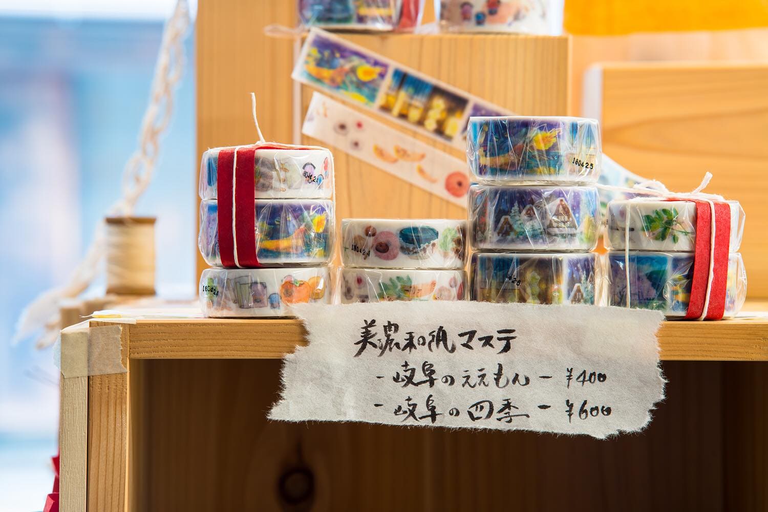 松久永助紙店オリジナルのマスキングテープが2種類、棚の上に並べられています。商品名は「岐阜のええもん」「岐阜の四季」です。1種類包装されたものと2種セットで包装されたものがあります。商品の近くには商品名と価格が記載された紙が貼ってあります。下記は関連するキーワードになります。美濃和紙 和紙 マスキングテープ 文具 特産品柄 四季柄 オリジナル ご当地 ならでは 岐阜 プレゼント お土産 プチギフト 松久永助紙店 近くで和紙が売ってるところ オンラインショップ
