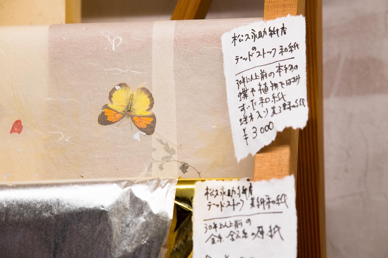 松久永助紙店の蔵出し和紙、本物の蝶入りの和紙がうつっています。30年以上前のデッドストック品、再生産ができない貴重な一品です。蝶入り和紙の下にはこちらも30年以上前の金糸、銀糸の原紙もうつっています。それぞれの和紙の横には商品名と価格、短い説明文がかかれた紙が貼ってあります。下記は関連するキーワードになります。蔵出し 蔵開き 珍しい 貴重 あるだけ 再生産できない 美濃和紙 和紙 レア デッドストック 蝶入り 植物入り 金糸 銀糸 松久永助紙店 近くで和紙が売ってるところ オンラインショップ