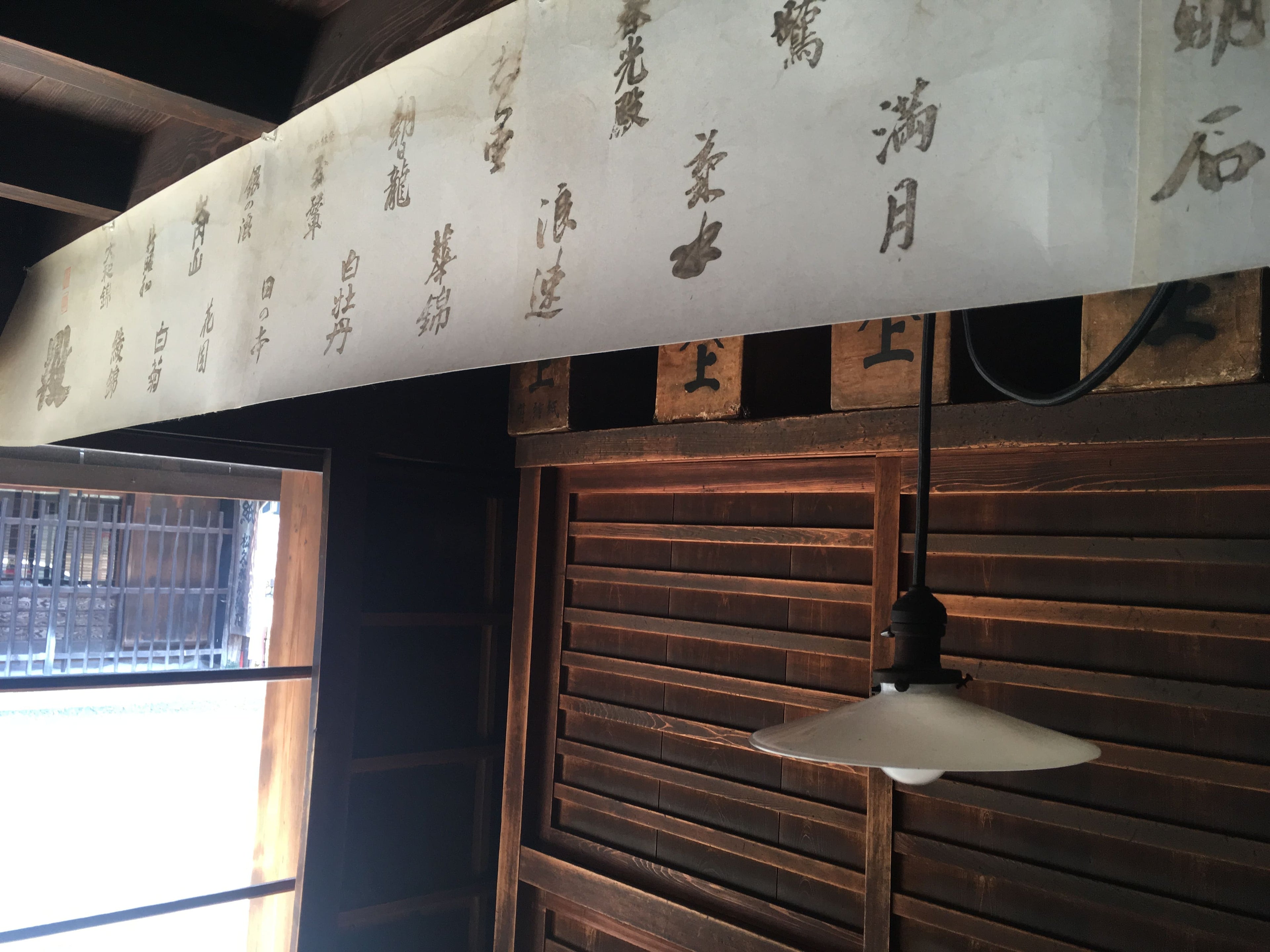 松久永助紙店の現店舗向かいにある帳場の中の様子がうつっています。壁に沿ってある箪笥、平笠シェードの電球、和紙でできた横断幕のようなものがうつっています。■松久永助紙店では紙糸(和紙糸)、紙布(和紙布)、美濃和紙靴下(和紙ソックス)、美濃和紙タオルなどの美濃和紙をつかった生活雑貨などを取り扱っています。主な商品は、和紙・美濃和紙(楮紙/こうぞ紙・マニラ麻・落水紙・本美濃紙/那須楮・墨流し)、和紙雑貨(カードケース・さついれ・干支カレンダー)、和紙文具(ノート・マスキングテープ・和紙はがき)、紙布雑貨(手ぬぐい・ワインバッグ・トートバッグ・ポーチ・ランチョンマット・マルチクロス)、生活雑貨(アクセサリー・ハンカチ・ハンドタオル・フェイスタオル・バスタオル・サウナタオル/ロングタオル・靴下/ソックス・アームカバー・信玄袋)、和紙布(帆布・ガーゼ・シャツ地・ニット・天竺・ヘリンボーン・手ぬぐい・コットン混・リネン混)、和紙糸(Kamito糸柱・ネロ・編み物用・織物用・刺繍用・玉売り・綛売り・コーン売り)です。