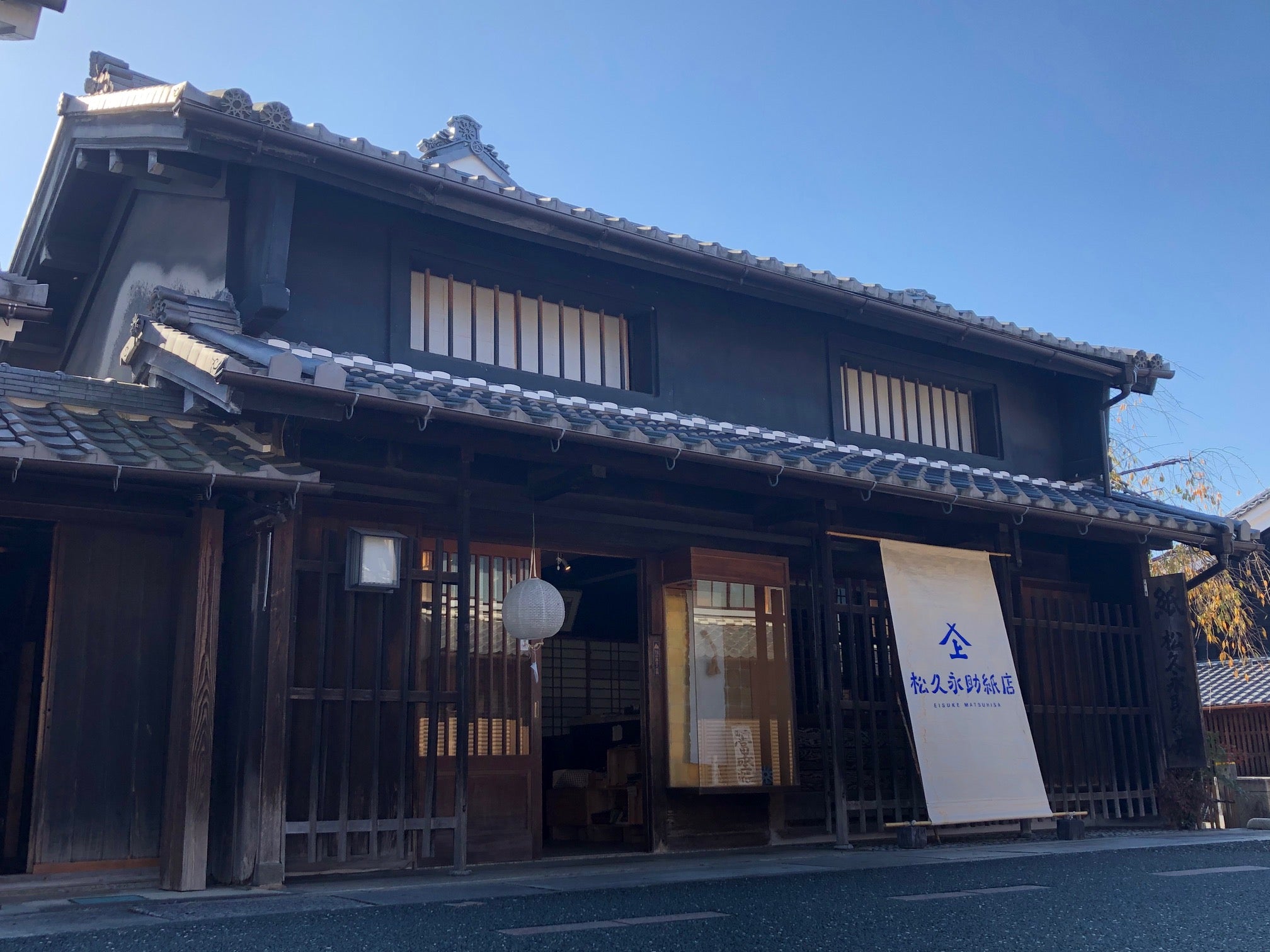 松久永助紙店の現店舗の外観がうつっています。2階建ての木造住宅で1階が店舗、店舗入り口には和紙の提灯がかけられ、右に少し離れた場所には、店名がはいった大きな暖簾があります。■松久永助紙店では紙糸(和紙糸)、紙布(和紙布)、美濃和紙靴下(和紙ソックス)、美濃和紙タオルなどの美濃和紙をつかった生活雑貨などを取り扱っています。主な商品は、和紙(楮紙/こうぞ紙・マニラ麻・落水紙・本美濃紙/那須楮・墨流し)、和紙雑貨(カードケース・さついれ・干支カレンダー)、和紙文具(ノート・マスキングテープ・和紙はがき)、紙布雑貨(手ぬぐい・ワインバッグ・トートバッグ・ポーチ・ランチョンマット・マルチクロス)、生活雑貨(アクセサリー・ハンカチ・ハンドタオル・フェイスタオル・バスタオル・サウナタオル/ロングタオル・靴下/ソックス・アームカバー・信玄袋)、和紙布(帆布・ガーゼ・シャツ地・ニット・天竺・ヘリンボーン・手ぬぐい・コットン混・リネン混)、和紙糸(Kamito糸柱・ネロ・編み物用・織物用・刺繍用・玉売り・綛売り・コーン売り)です。