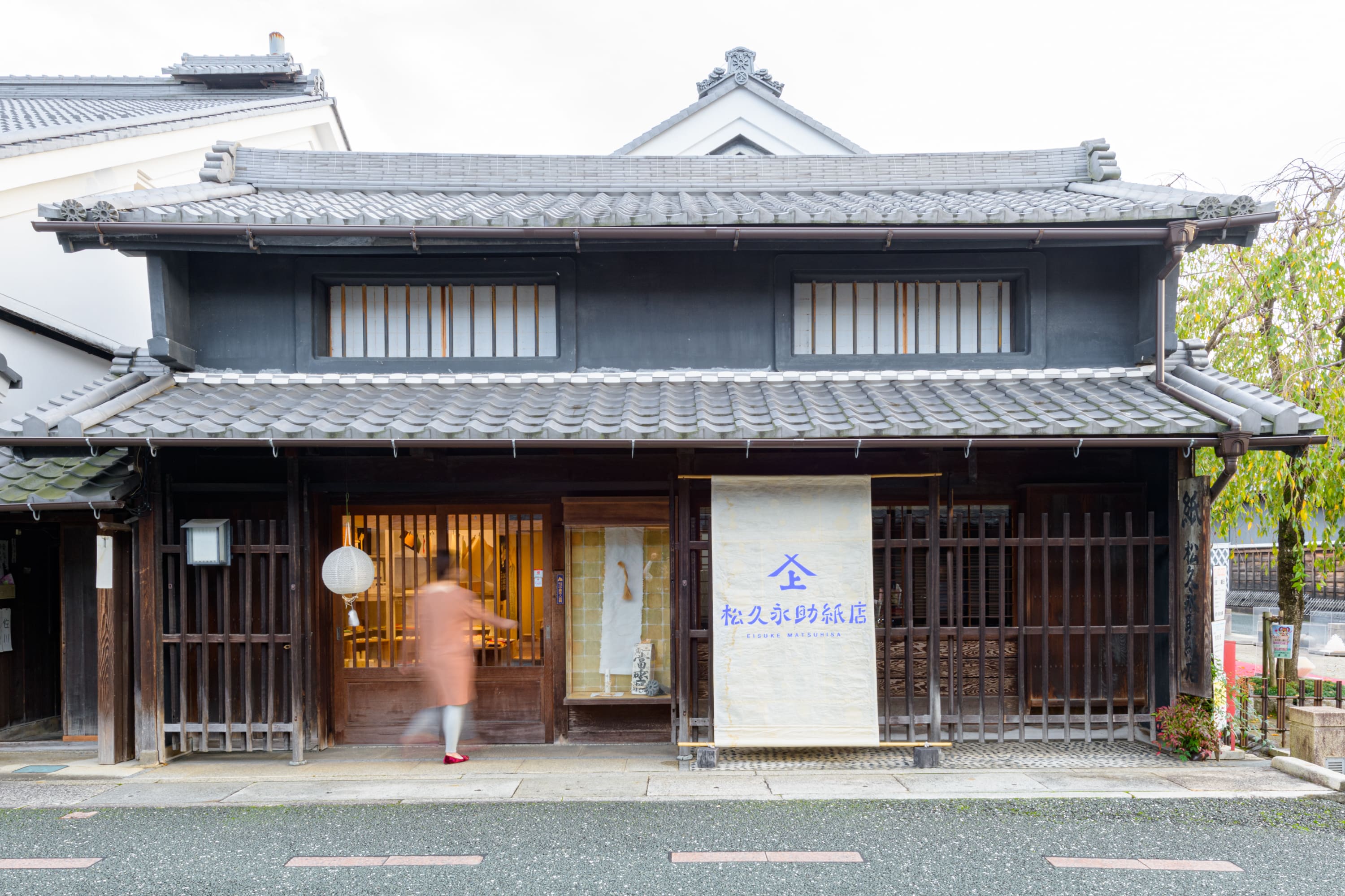 松久永助紙店の現店舗を正面からとった様子です。女性が店舗入り口の扉を開けようとしています。扉近くの軒先には和紙でできた提灯がかかっています。扉の右側にはガラス張りのショーケースと、そのまた右には店名がはいった大きなのれんがあります。■松久永助紙店では紙糸(和紙糸)、紙布(和紙布)、美濃和紙靴下(和紙ソックス)、美濃和紙タオルなどの美濃和紙をつかった生活雑貨などを取り扱っています。主な商品は、和紙・美濃和紙(楮紙/こうぞ紙・マニラ麻・落水紙・本美濃紙/那須楮・墨流し)、和紙雑貨(カードケース・さついれ・干支カレンダー)、和紙文具(ノート・マスキングテープ・和紙はがき)、紙布雑貨(手ぬぐい・ワインバッグ・トートバッグ・ポーチ・ランチョンマット・マルチクロス)、生活雑貨(アクセサリー・ハンカチ・ハンドタオル・フェイスタオル・バスタオル・サウナタオル/ロングタオル・靴下/ソックス・アームカバー・信玄袋)、和紙布(帆布・ガーゼ・シャツ地・ニット・天竺・ヘリンボーン・手ぬぐい・コットン混・リネン混)、和紙糸(Kamito糸柱・ネロ・編み物用・織物用・刺繍用・玉売り・綛売り・コーン売り)です。