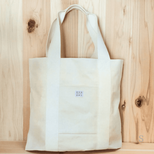 木目の背景に、和紙布でできたトートバッグがうつされています。色は生成りです。バッグ真ん中には店名が記載されたタグがついています。下記は関連するキーワードになります。紙布 和紙布 自然素材 11号帆布 軽い 紙糸 和紙糸 オリジナル コラボ商品 Sifuあだちや トートバッグ シンプル 使いやすい 松久永助紙店 オンラインショップ