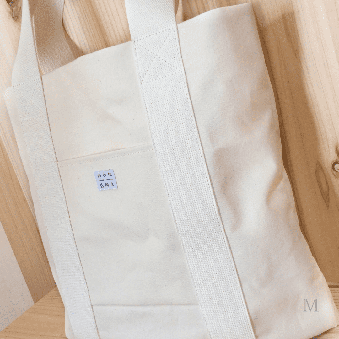 和紙布でできたトートバッグを右斜めから、やや寄り気味で撮影した写真です。色は生成りです。バッグ真ん中にはポケットと店名が記載されたタグがついています。下記は関連するキーワードになります。紙布 和紙布 自然素材 11号帆布 軽い 紙糸 和紙糸 オリジナル コラボ商品 Sifuあだちや トートバッグ 軽量バッグ シンプル 使いやすい カジュアル プレゼント 松久永助紙店 オンラインショップ