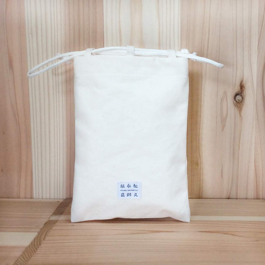 木目の背景に、和紙布でできた信玄袋がうつされています。色は生成り、下部には店名が記載されたタグがついています。袋の口は紙糸の組紐で締められる巾着式になっています。下記は関連するキーワードになります。紙布 和紙布 自然素材 11号帆布 軽い 紙糸 和紙糸 オリジナル コラボ商品 Sifuあだちや 信玄袋 巾着 シンプル 使いやすい 伝統的 日本製 こだわり プレゼント おすすめ 松久永助紙店 オンラインショップ