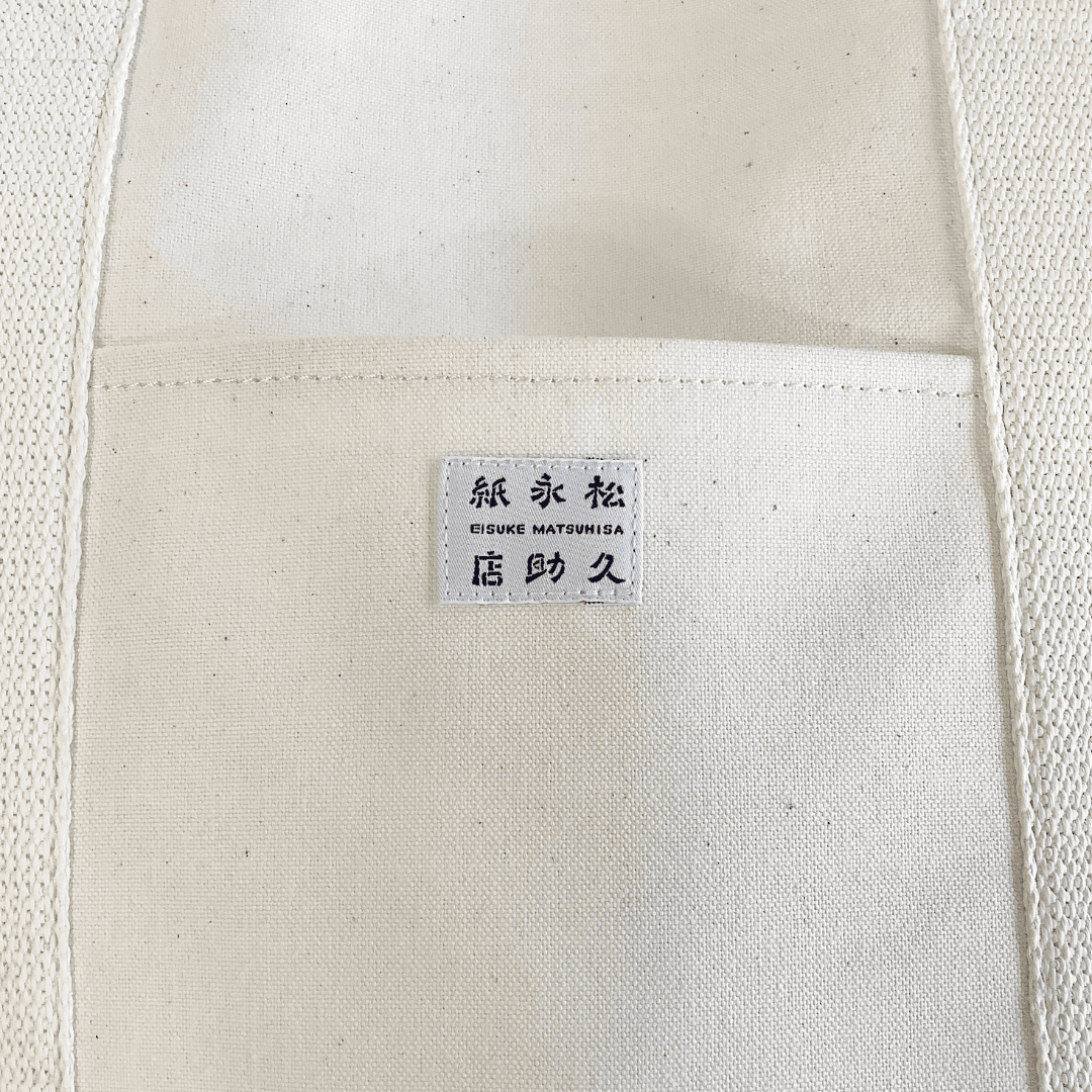 和紙布でできたトートバッグの外ポケットをアップで撮影した写真です。色は生成りで、真ん中には店名が記載されたタグがついています。下記は関連するキーワードになります。ポケットあり トートバッグ 軽量バッグ 普段使いしやすい 合わせやすい マチあり 紙糸 紙布 日本製 上質 こだわり 松久永助紙店 オンラインショップ