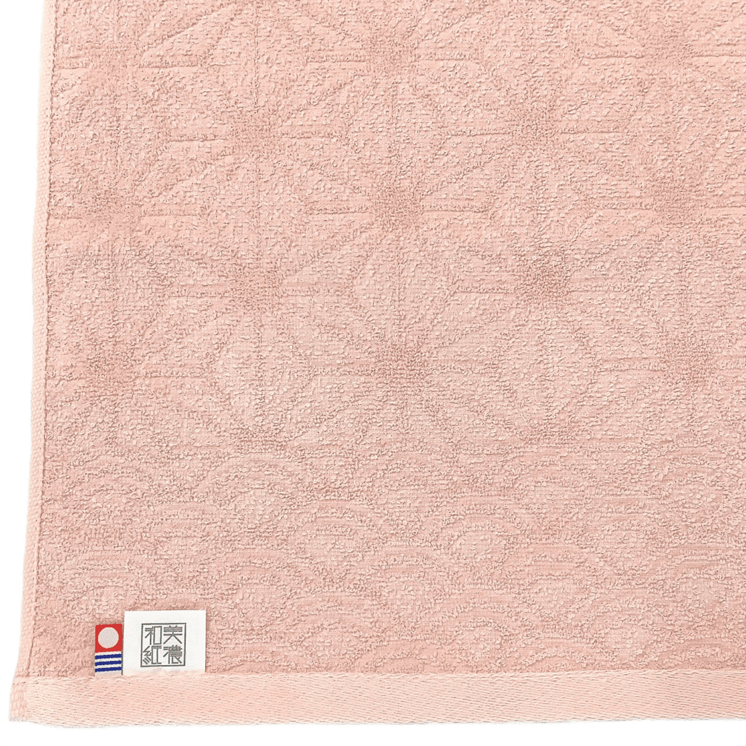 美濃和紙タオル、花麻柄、桜色、タグが付いているところがアップでうつっています。タグは「美濃和紙」とかかれたもの（右側）と、今治タオルのブランドロゴ（左側）の2つがついています。下記は関連するキーワードになります。和紙タオル ハンドタオル フェイスタオル 美濃和紙 機能的 吸水性 軽い シャリ感 経年変化 和柄 お祝い 引き出物 ピンク 推しカラー パステルカラー プレゼント ギフト ご挨拶 松久永助紙店 オンラインショップ
